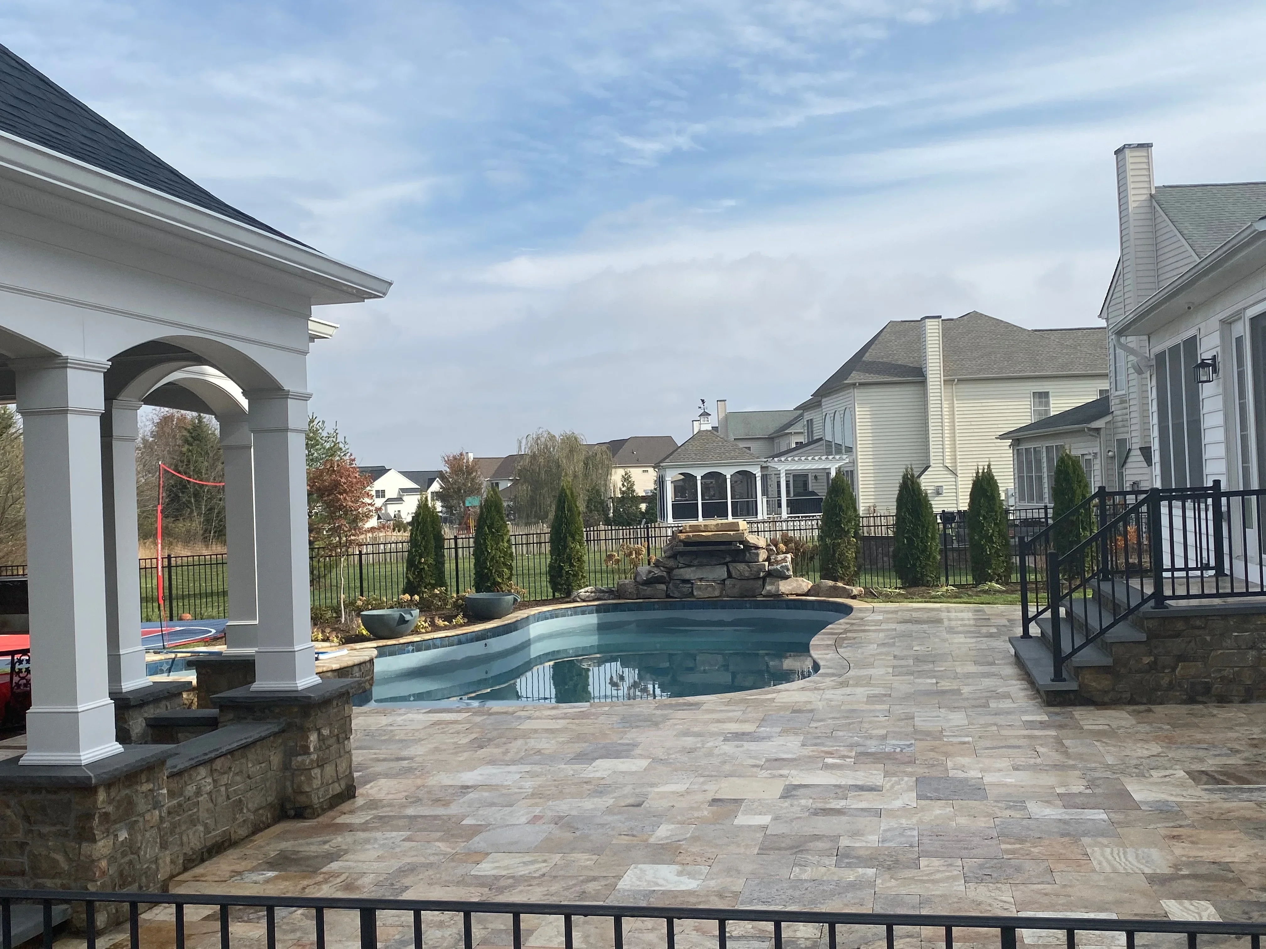 Stone paver patio with swimming pool, white pergola, and black aluminum fencing in residential backyard