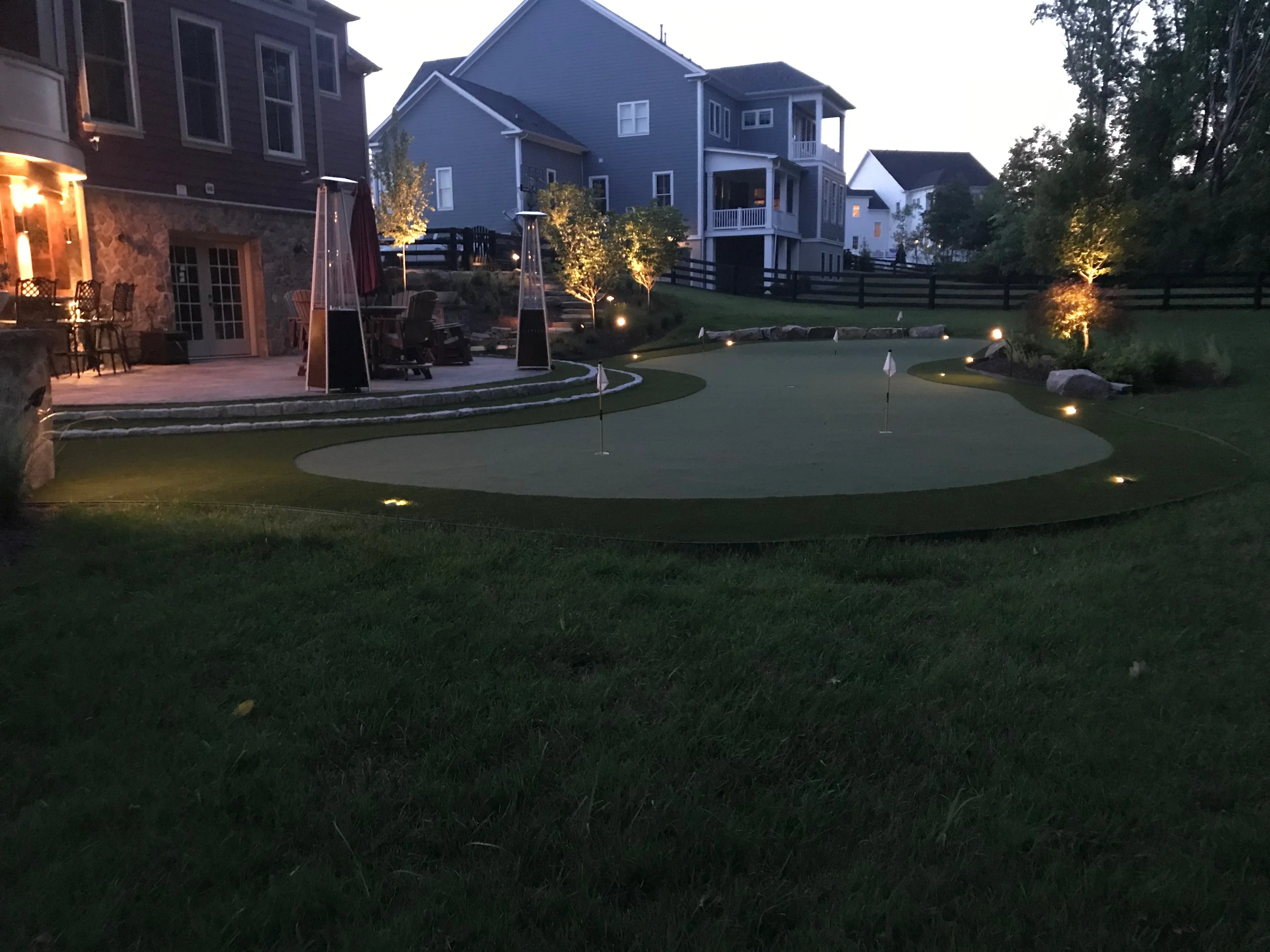 Synthetic putting green with integrated landscape lighting, curved edges, and flagsticks at dusk
