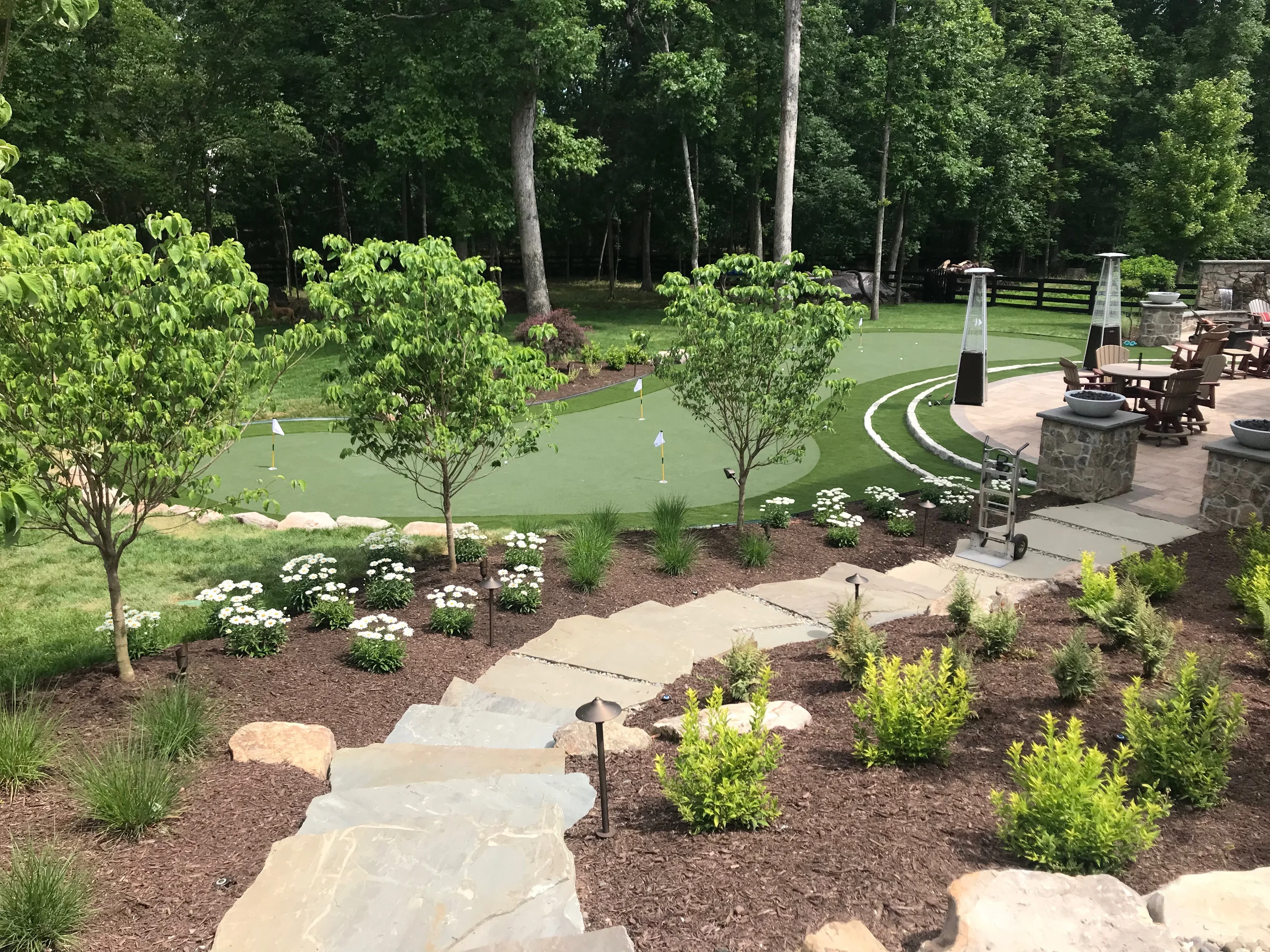 Stone patio with curved pathways, putting green, landscape beds with shrubs and flowers, and integrated outdoor seating area