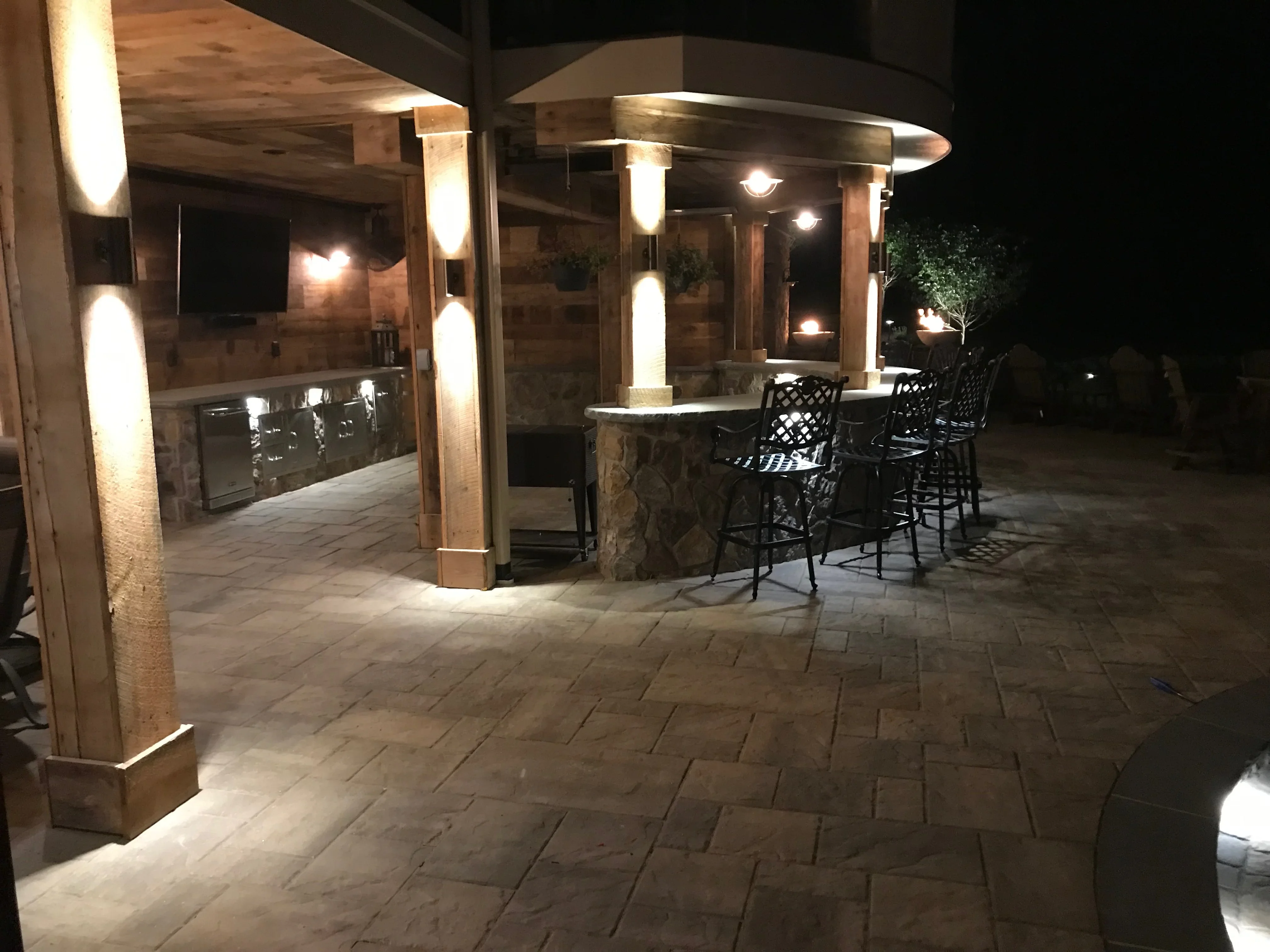 Lit patio with pergola, wooden posts, stone bar counter, and paver surface at night