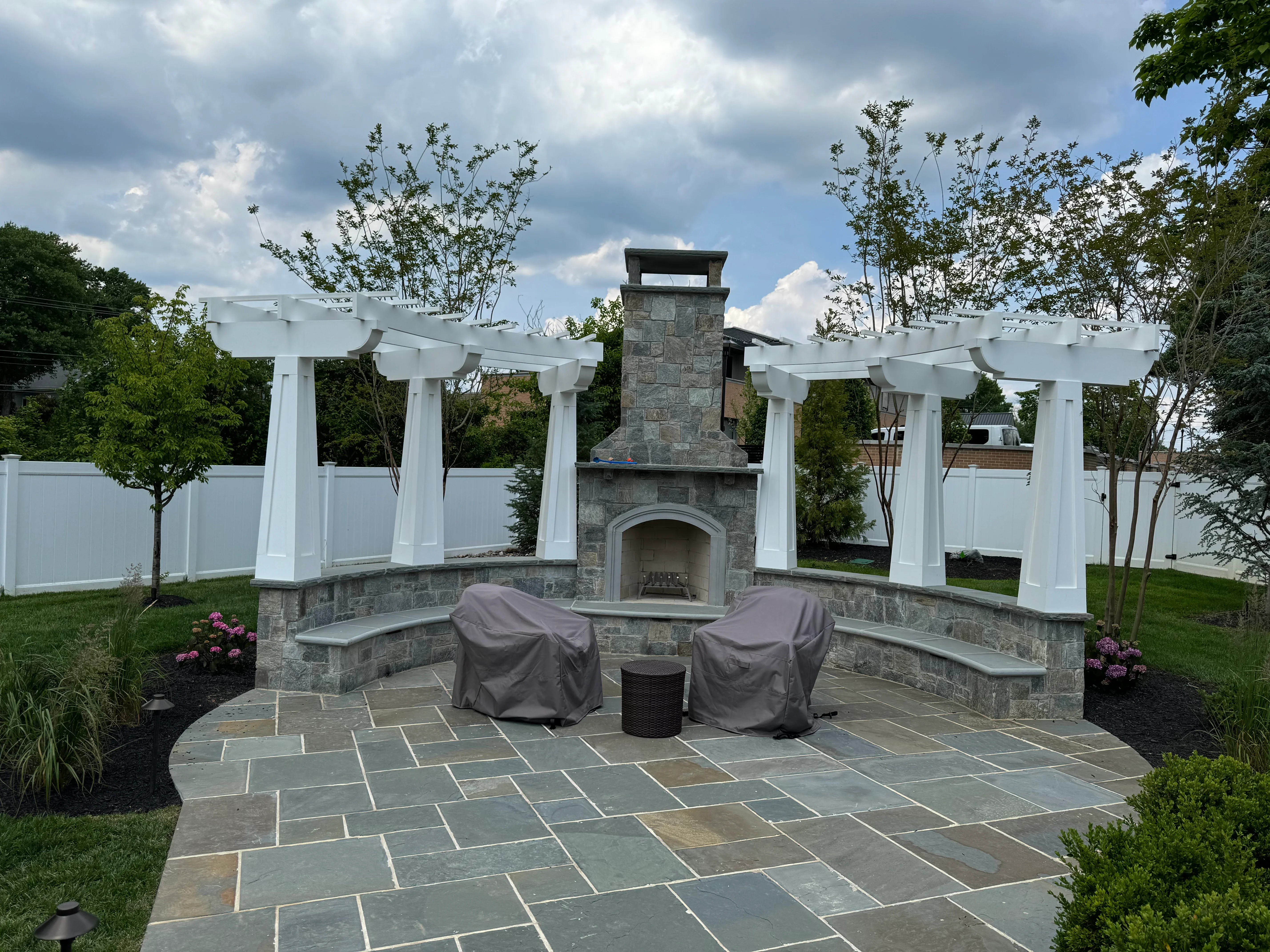 Flagstone patio with stone fireplace, white pergola, curved benches, and landscaping